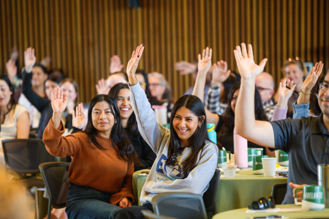 Cal Poly employees sitting at a presentation, raising their hands and smiling. 