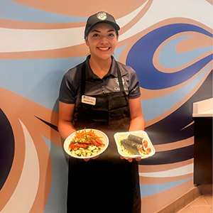 A Campus Dining student associate holds two Kai Poke menu items beneath Kai Poke signage inside 1901 Marketplace.