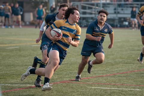 Zion Jerge carrying a rugby ball down the pitch with other players in pursuit.