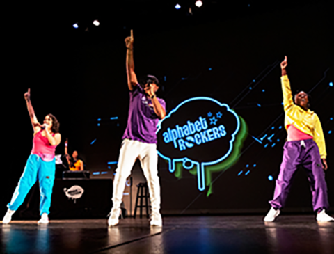 Alphabet Rockers performing on stage with a neon sign and a DJ behind them.