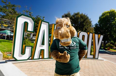 Musty Mustang giving a thumbs up while standing in front of the Cal Poly sign.