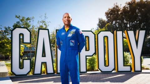 Victor Glover in a blue NASA jumpsuit standing in front of the Cal Poly sign.