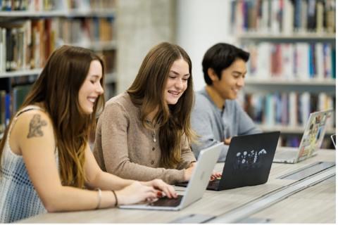 Students working on laptops at a table with shelves of books in the background.