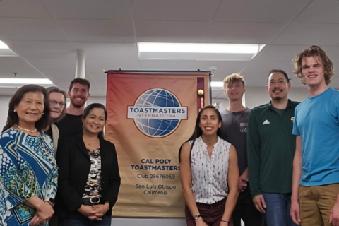 Members of Cal Poly Toastmasters posing for a photo next to an event sign.