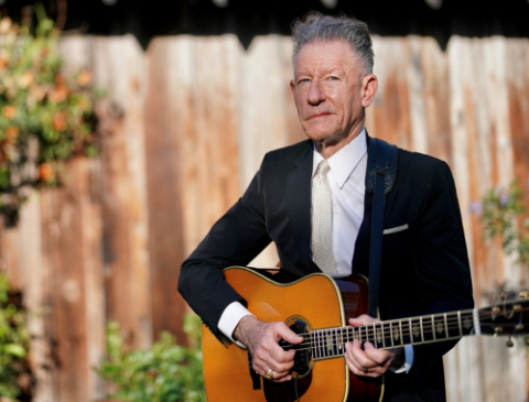 Lyle Lovett in a suit holding his guitar with a fence and plants in the background.