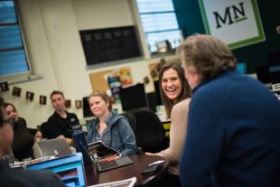A group of students around a table talking and laughing with an advisor in the Mustang Media Group conference room