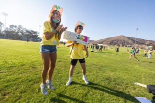 Two students in yellow Week of Welcome T-shirts smiling and holding up a sign reading Group 162 on a grassy field