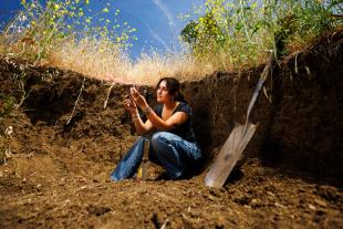 A student sits in a dirt pit with flowering grasses at the top. They examine some soil.