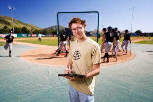 Gabe Riedel holds a clipboard on the baseball field with players lined up at bat behind him.