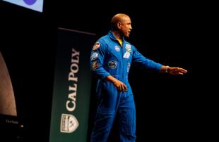 A man in a blue NASA jumpsuit speaks onstage in front of a Cal Poly banner