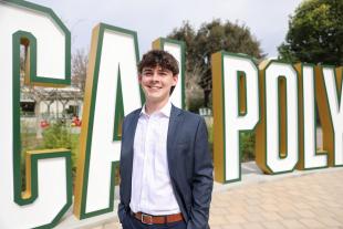 A young man in a suit stands in front of a large Cal Poly sign.