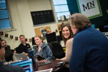 A group of students around a table talking and laughing with an advisor in the Mustang Media Group conference room