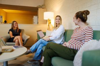 A student sits on a green couch in between two older staff members. The three smile as the student shows them her computer screen.