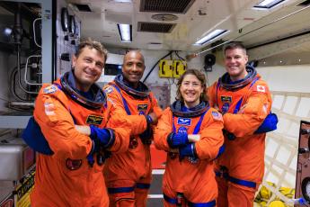 Artemis II NASA astronauts (left to right) Reid Wiseman, Victor Glover, and Christina Koch, and CSA (Canadian Space Agency) astronaut Jeremy Hansen wear orange suits and stand in a room at Kennedy Space Center.