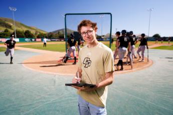 Gabe Riedel holds a clipboard on the baseball field with players lined up at bat behind him.