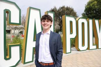 A young man in a suit stands in front of a large Cal Poly sign.