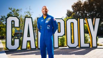 Victor Glover wears a blue NASA flight suit standing in front of the Cal Poly letters on campus