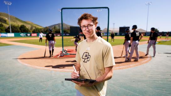 A statistics student holding a clipboard and standing on a baseball field in front of baseball players.