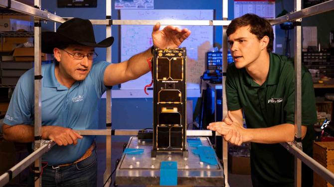 A professor in a blue shirt and cowboy hat inspects a CubeSat while a student in a green shirt looks on.