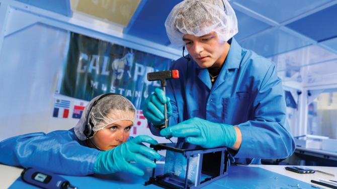 Two students in hair nets and blue coats work on a small satellite in a clean room