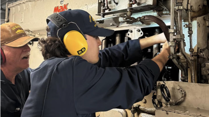 A cadet wearing ear protection getting instruction from an instructor in the engine room.