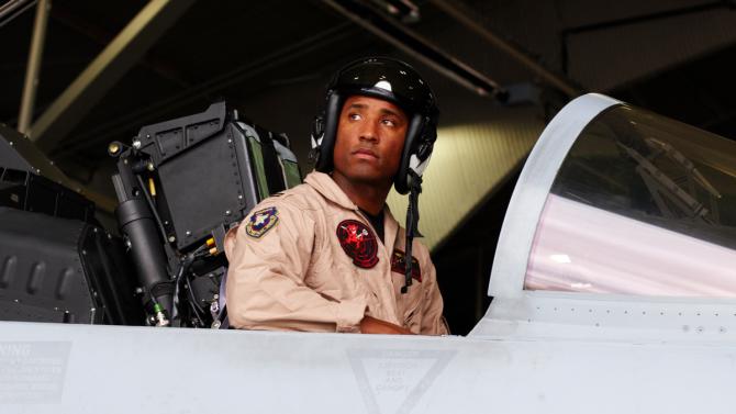 Victor Glover wears a helmet and tan flight suit while sitting in the cockpit of a fighter jet