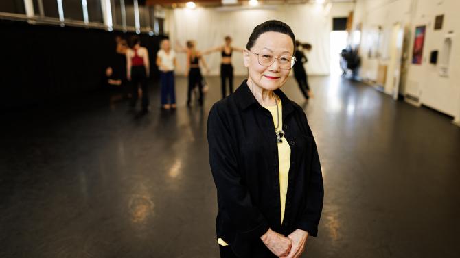 A woman in a black jacket stands in front of dancers rehearsing in a studio