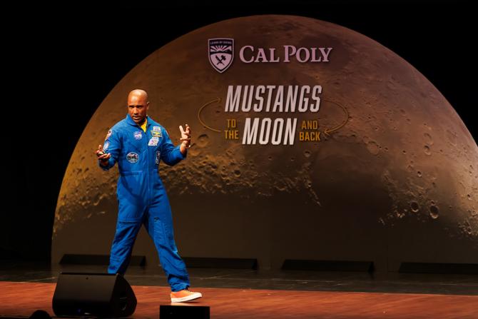A man in a blue NASA jumpsuit speaks onstage in front of an image of the moon with the words "Cal Poly - Mustangs to the Moon and back"