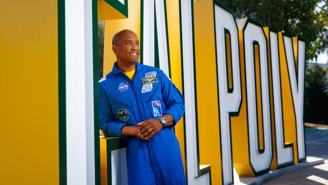 Victor Glover in a space jumpsuit leaning against the Cal Poly sign.
